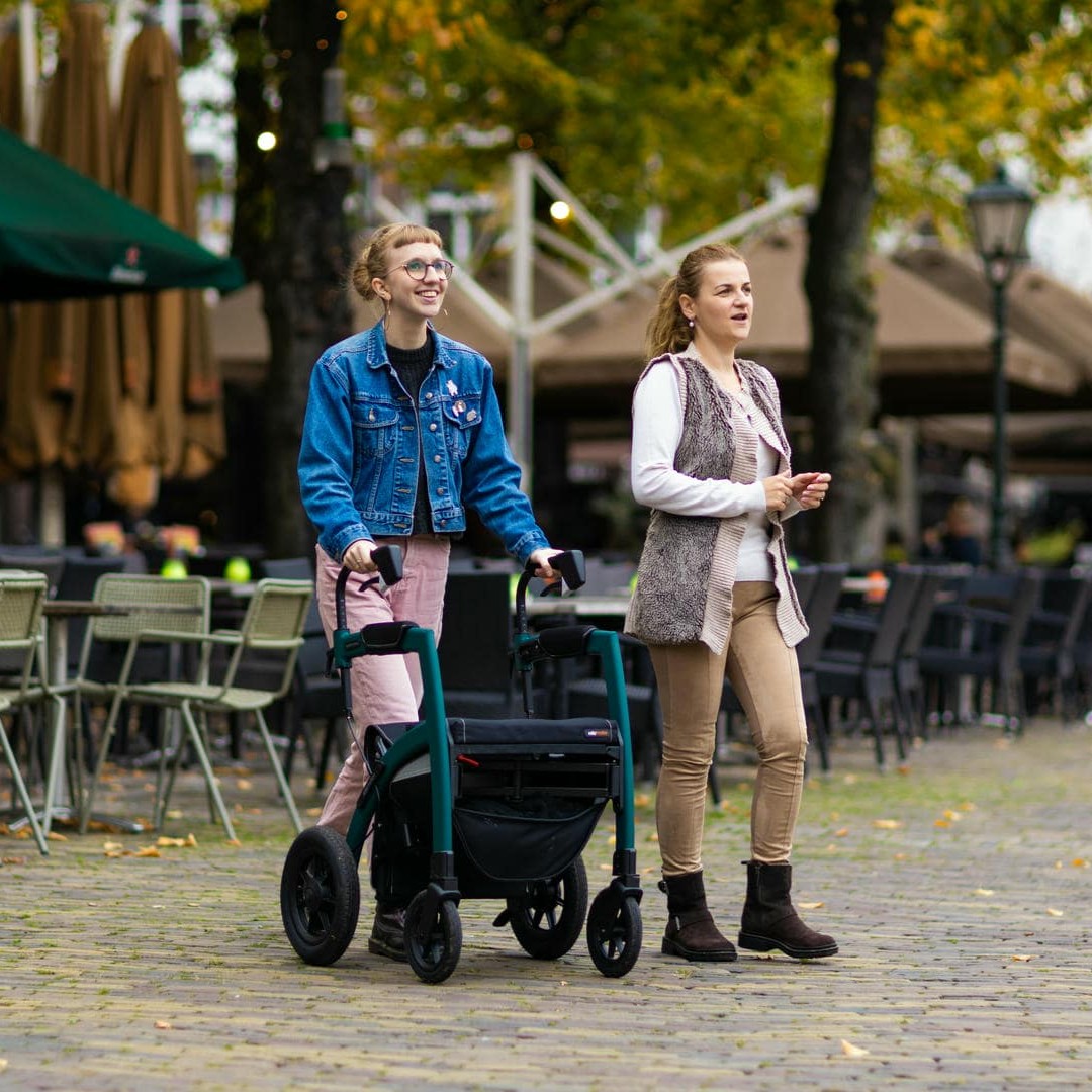 La photo montre deux femmes, dont l’une avec un déambulateur, sur une place publique entourée de cafés et d'arbres.