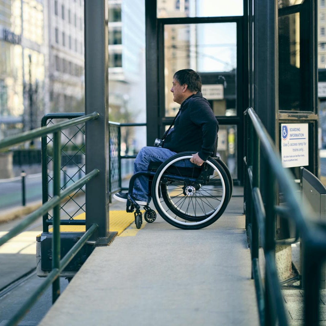 La photo montre un homme en fauteuil roulant qui attend un moyen de transport public près d'une rampe.