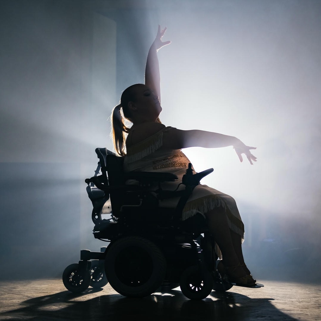 La photo montre une femme en fauteuil roulant, les bras artistiquement écartés vers le haut et devant. Elle est sur une scène et la lumière des projecteurs semble telle que l'on ne voit que son relief.