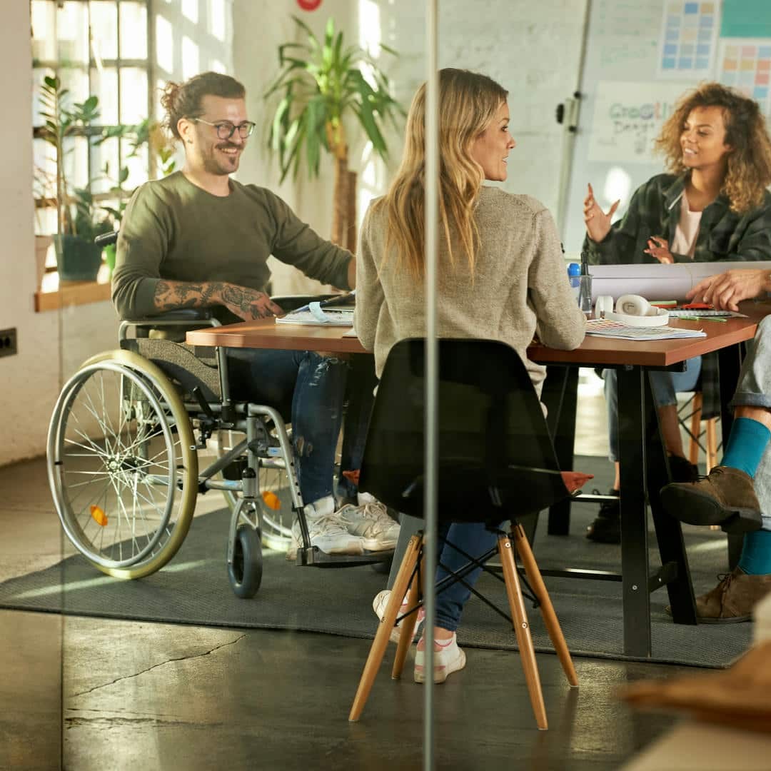 La photo montre quatre personnes autour d'une table. L'une d'entre elles est assise dans un fauteuil roulant. En arrière-plan, un tableau avec de nombreux documents. L'atmosphère semble détendue, tout le monde rit.