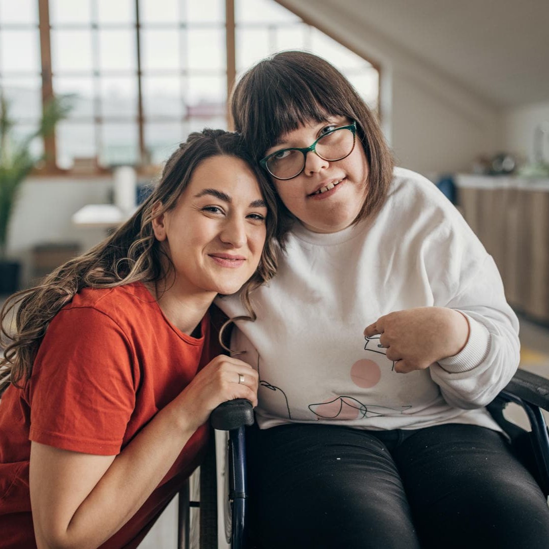 La photo montre une jeune fille en fauteuil roulant et une jeune femme agenouillée à ses côtés. Toutes deux ont un bras enroulé autour des épaules de l'autre et sourient.