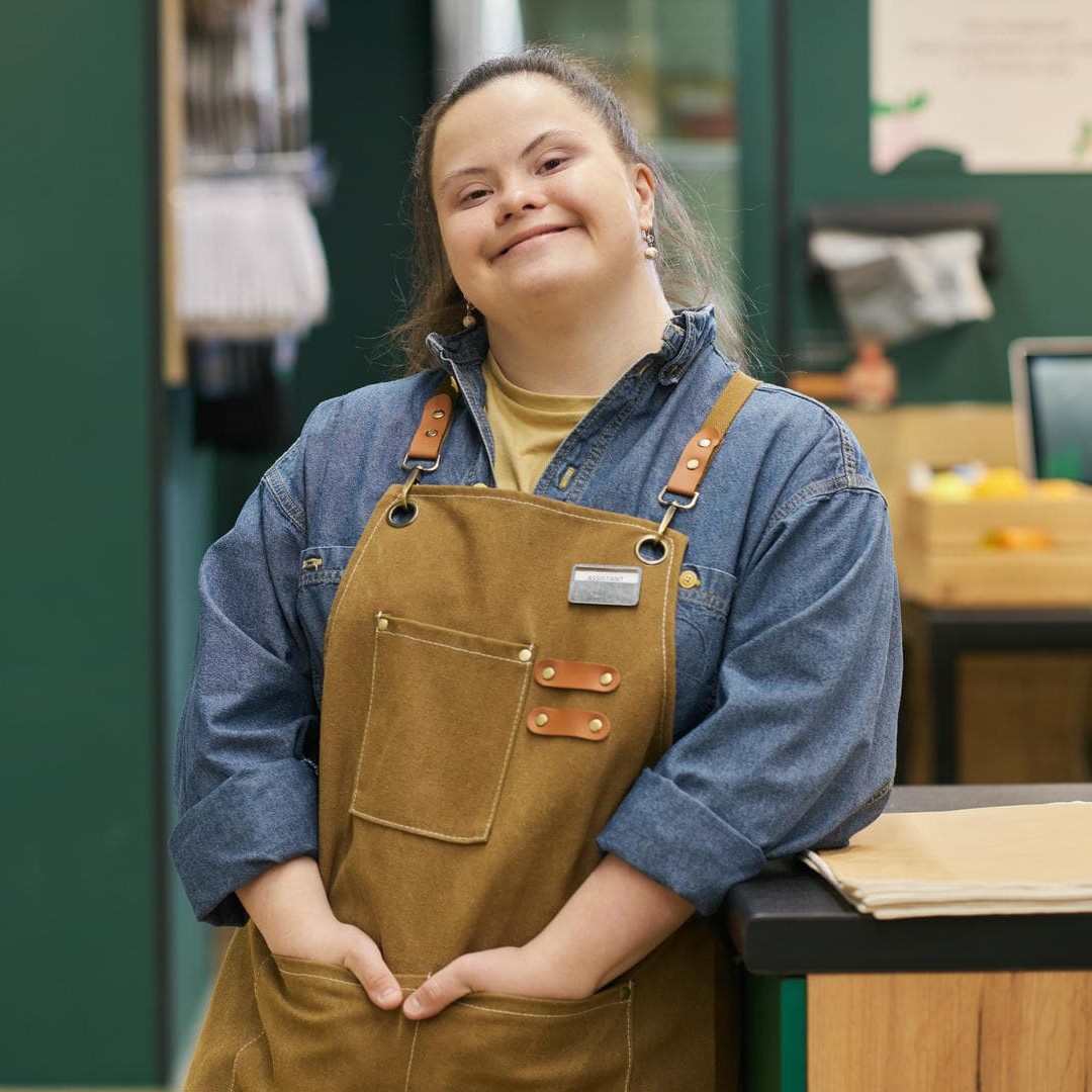 Das Foto zeigt eine hübsche, junge Frau mit Down-Syndrom, die in Arbeitskleidung in einem Warenlager steht.