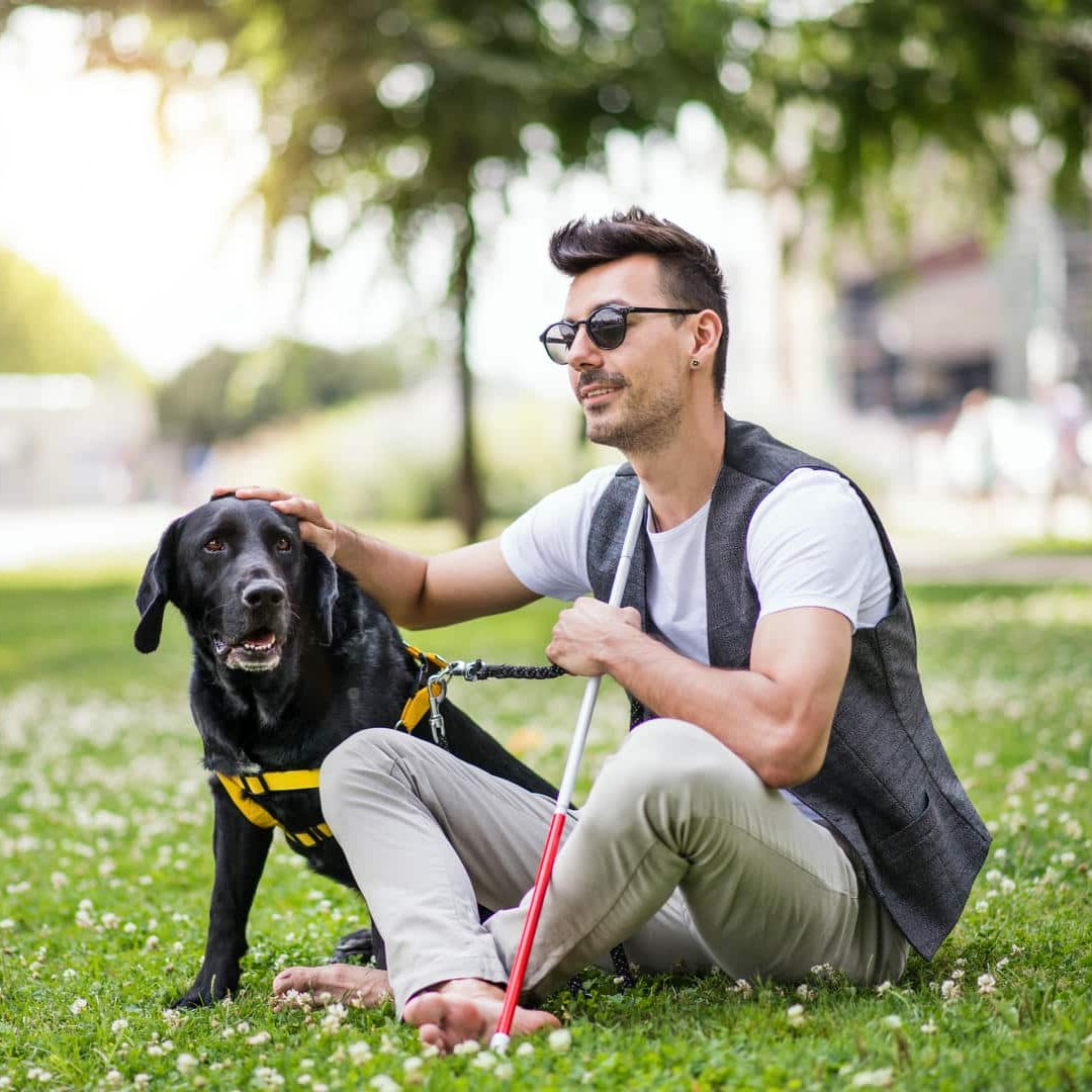 La photo montre un jeune homme portant des lunettes de soleil et une canne blanche, assis sur la pelouse d'un parc à côté de son chien d'assistance.