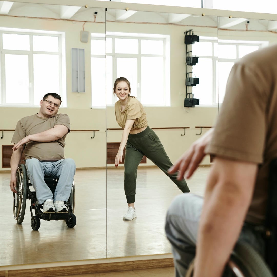 La photo montre deux personnes devant des miroirs dans une salle de danse. Une jeune femme se tient à côté d'un homme en fauteuil roulant, tous deux dansent et étendent leur bras droit vers la gauche.