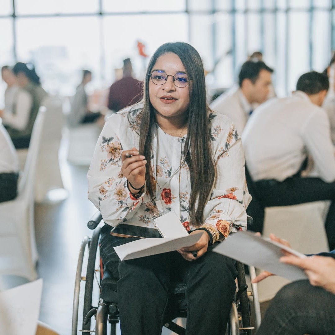 La photo montre une jeune femme en fauteuil roulant dans une conférence, entourée d'un groupe de personnes. Elle tient un stylo et une feuille de papier et s'apprête à discuter.