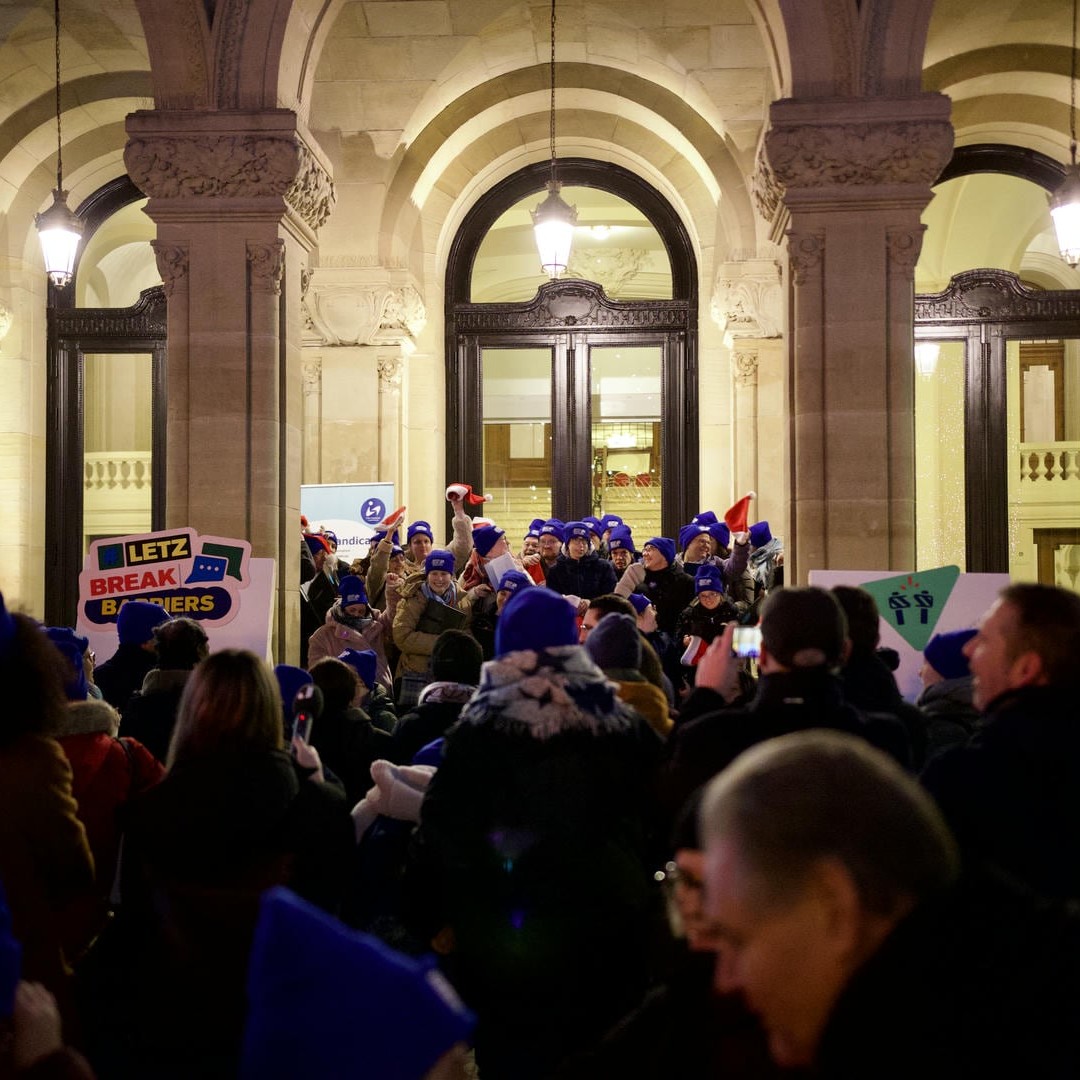 La photo montre une assemblée de personnes écoutant attentivement un groupe portant des casquettes bleues devant le Cercle Cité dans la Ville de Luxembourg. Il s’agit de l’événement #LetzBreakBarriers 2024.