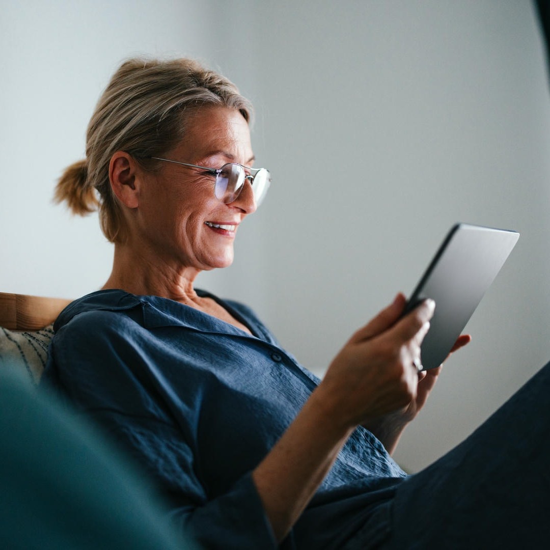 La photo montre une femme souriante, allongée sur une chaise confortable, qui regarde une tablette.