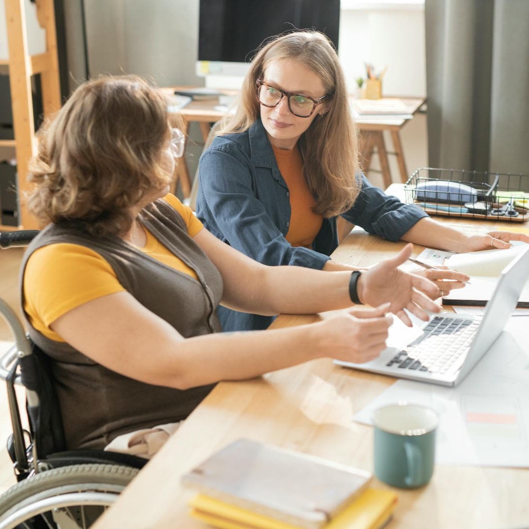 Das Foto zeigt zwei Frauen, die nebeneinander an einem Tisch sitzen, eine davon im Rollstuhl. Vor sich haben sie einen Laptop und sie diskutieren angeregt.