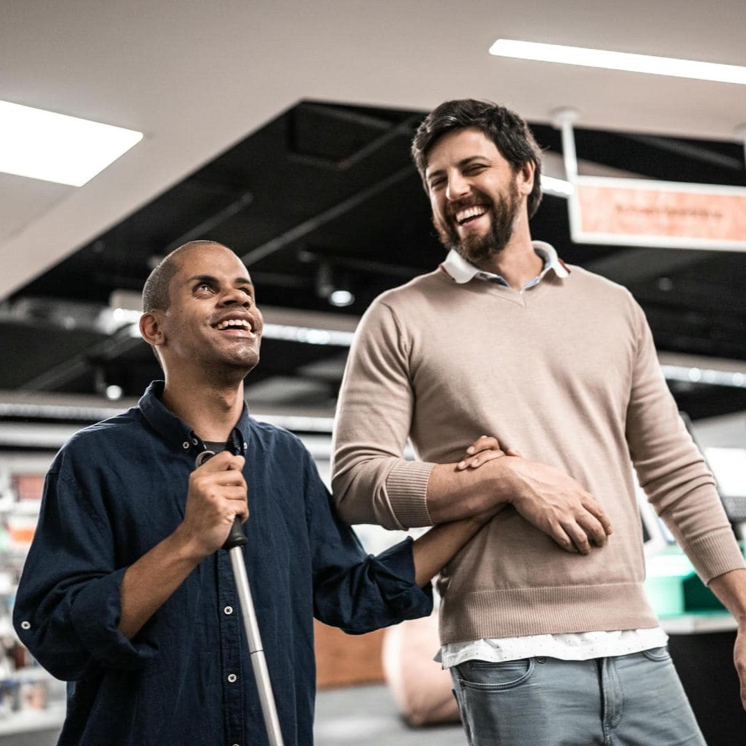 Das Foto zeigt zwei Männer; einer davon ist blind und wird von dem anderen geführt. Beide lachen herzhaft.