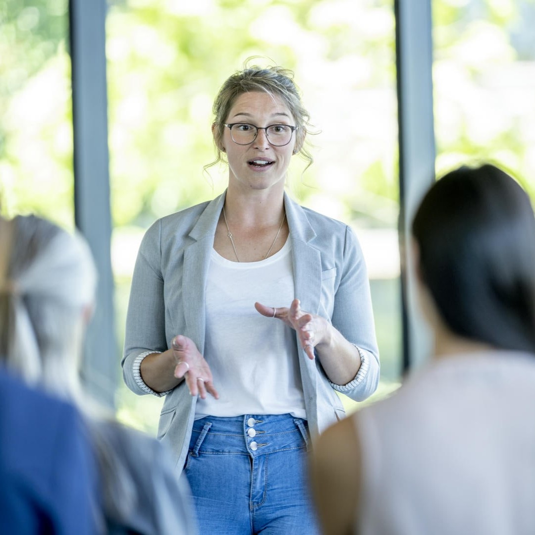 Das Foto zeigt eine junge Frau vor einem Publikum. Sie wirkt freundlich und dynamisch, hinter ihr sind Fenster, durch die die Sonne scheint.