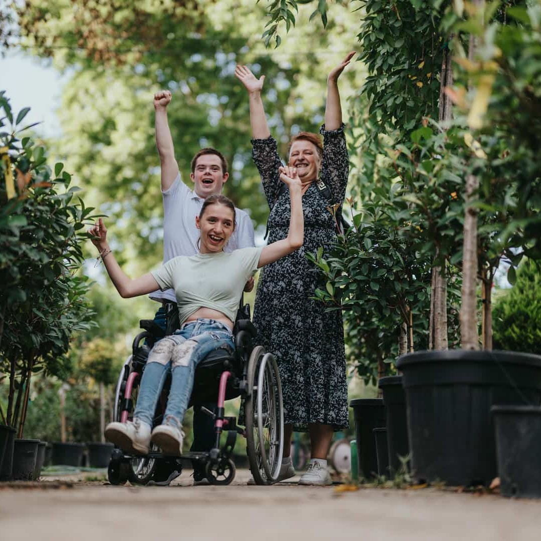 La photo montre trois personnes jetant joyeusement leurs mains en l'air : une femme d'âge moyen, un jeune homme et une adolescente en fauteuil roulant.