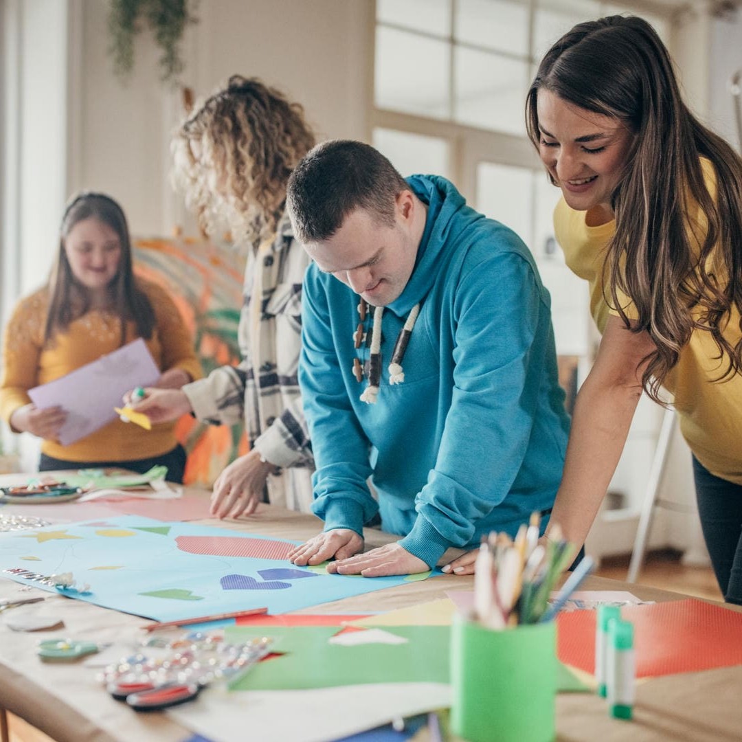 La photo montre un groupe de personnes avec et sans handicap visible, réunies autour d'une table avec du matériel de bricolage. Elles travaillent ensemble à un projet créatif.