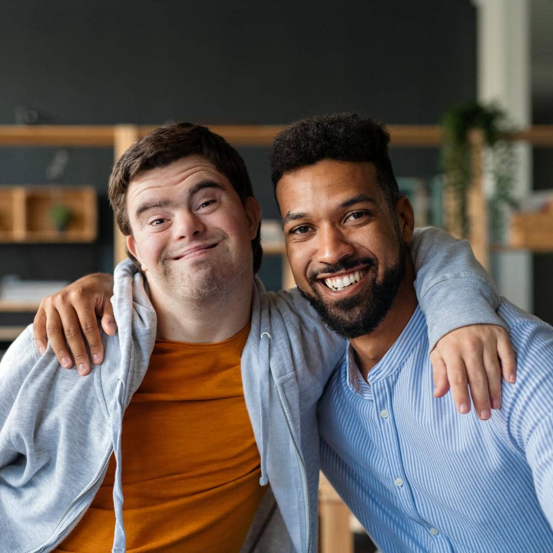 Un jeune homme atteint d’une trisomie 21 et un jeune homme sans handicap visible sont assis côte à côte, chacun ayant un bras enroulé autour de l'épaule de l'autre. Tous deux sourient.