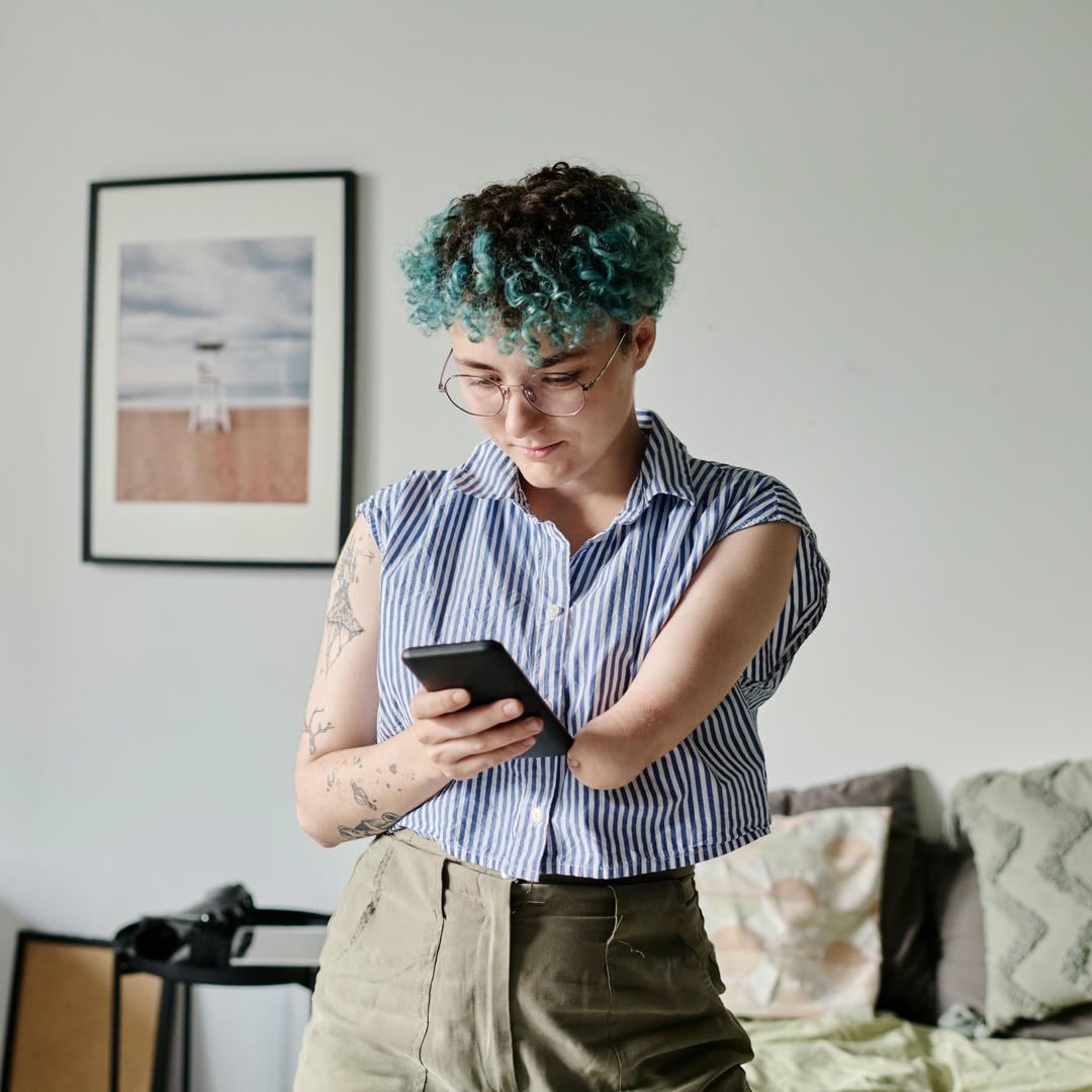 La photo montre une jeune femme aux cheveux bleus et frisés qui regarde un téléphone portable avec concentration. Elle n'a qu'une seule main.