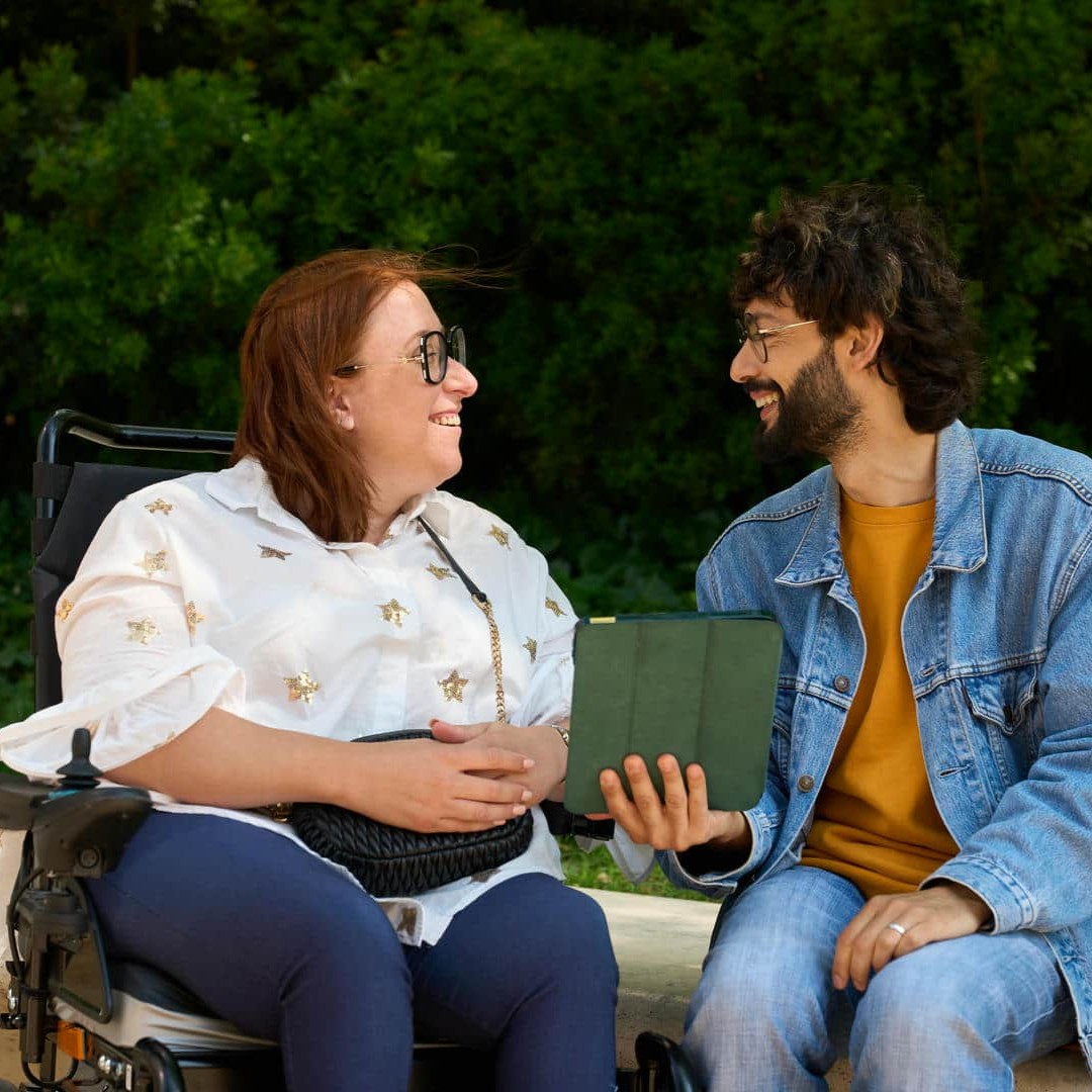 La photo montre un jeune homme assis sur un mur à côté d'une femme en fauteuil roulant. Il tient une tablette à la main et tous deux parlent et sourient.