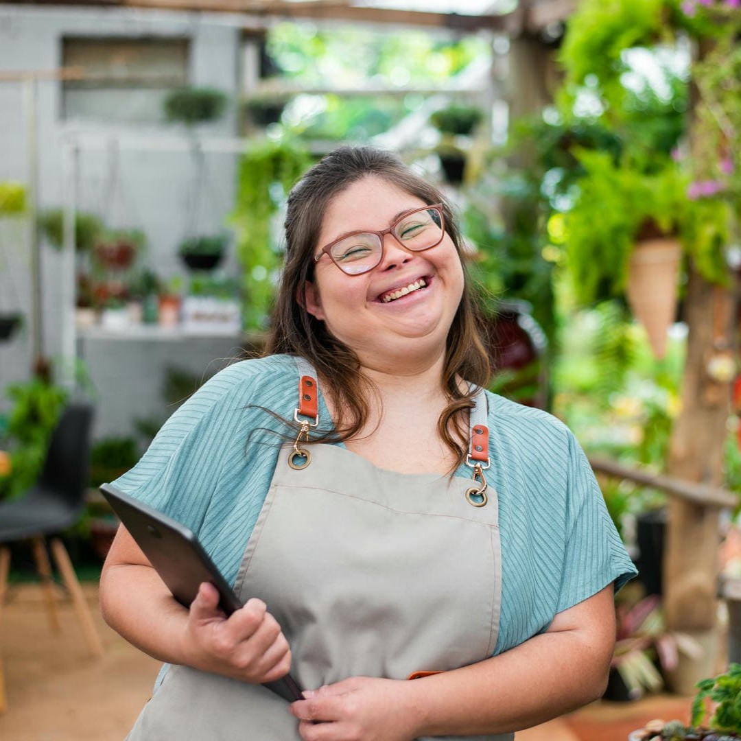 La photo montre une jeune femme avec trisomie 21 souriante. Elle porte un tablier et est entourée d'étagères avec des plantes.