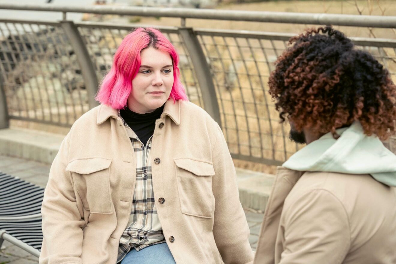Zwei Personen sprechen miteinander. Von vorne, eine Frau mit rosa Haaren. Von hinten, ein Mann mit dunklen, lockigen Haaren.