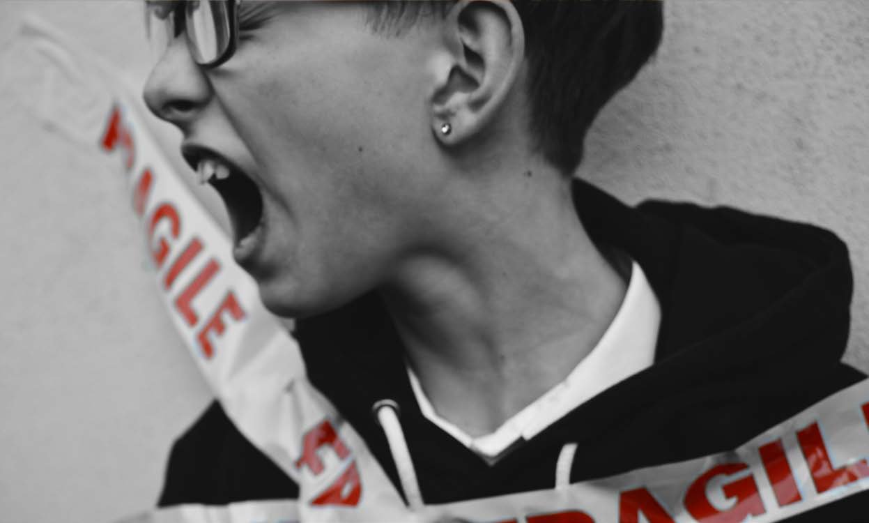 Schwarz-Wei&szlig;-Foto: Eine Person, die von der Seite zu sehen ist, schreit. Um sie herum ist ein rot-wei&szlig;es Band mit der Aufschrift &bdquo;FRAGILE&ldquo; gewickelt.
