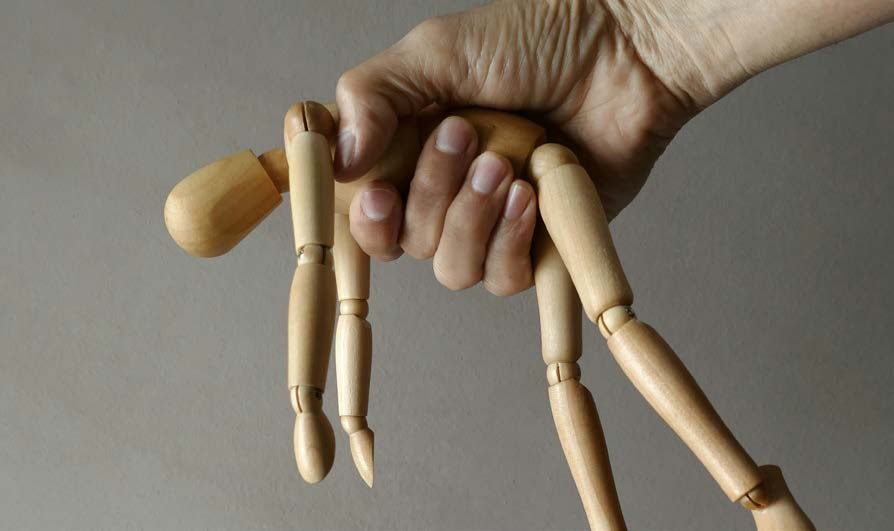 Eine Hand h&auml;lt eine h&ouml;lzerne Gliederpuppe fest. Die Puppe liegt in der Hand wie ein lebloser K&ouml;rper.