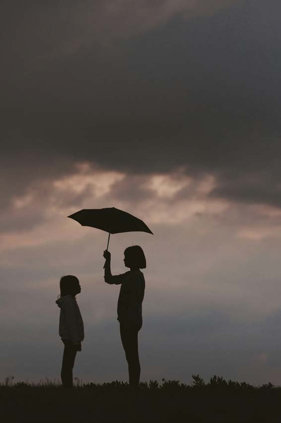 Silhouetten in der D&auml;mmerung: Ein Erwachsener h&auml;lt einen Regenschirm &uuml;ber ein Kind, unter bew&ouml;lktem Himmel.